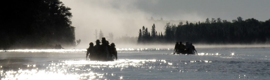 Voyageurs paddle down a wide, winding river in 19th century style canoes and attire